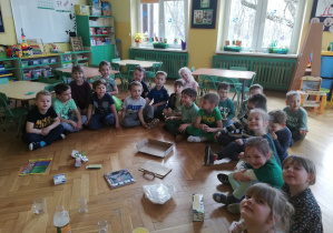 Grupa przedszkolaków siedzi na podłodze, przed dziećmi na podłodze znajdują się surowce wtórne : papier, plastik, szkło.
